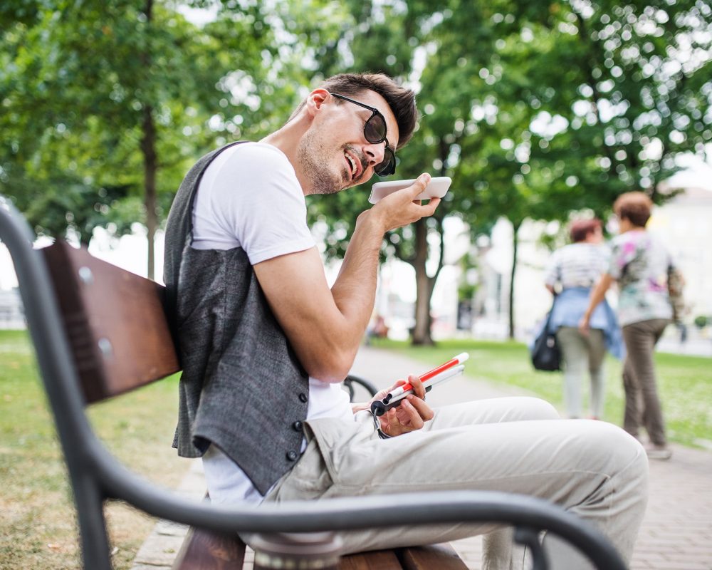 A young blind man with smartphone sitting on bench in park in city.