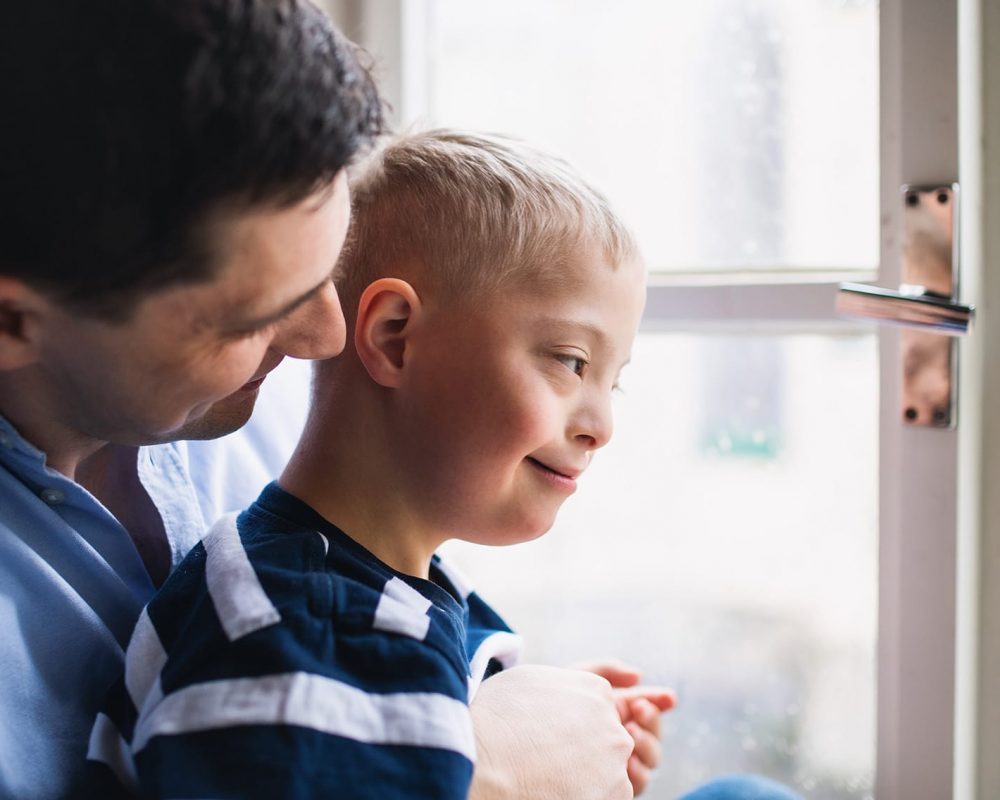 Mature father with happy down syndrome son indoors at home, looking through window.